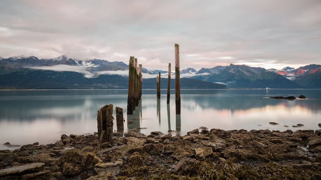Calm Water Mountains Wooden Posts Rocky Shore Mist Clouds