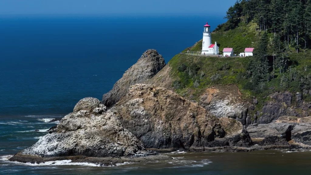 lighthouse ocean cliff rocky greenery buildings blue sky waves