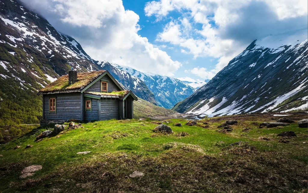Mountainous Landscape Wooden Cabin Snow-Capped Peaks Green Grass Rocky Terrain Blue Sky